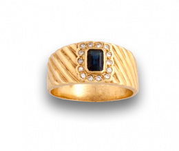 71.  Sortija con zafiro cuadrado orlado de brillantes en montura ancha gallonada de oro amarillo de 18K.