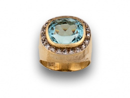 99.  Sortija con montura elevada y aguamarina de talla oval orlada de brillantes en oro amarillo de 18K.